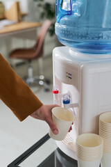Caucasian young adult hand filling paper cup with water from office water cooler, disposable cups stacked nearby, modern workspace background visible in soft focus