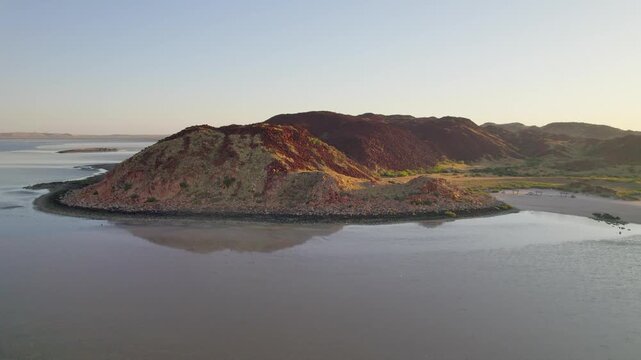 Aerial drone view of a rocky coastal hill near Karratha on the Dampier Peninsula. Calm water surrounds the landform as warm light reveals textures, curves and the remote Western Australian coastline.
