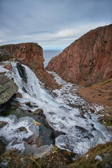 Water flows down a rocky cliff into a frozen river in Teriberka, Russia during the winter months with cloudy skies above