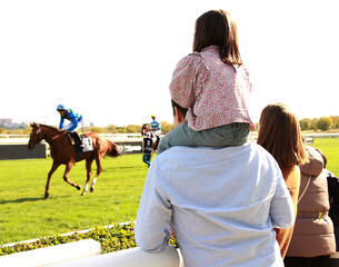 Familia de p&uacute;blico en hip&oacute;dromo con ni&ntilde;a a hombros a caballlito de espectador disfrutando plan de ocio en domingo de fin de semana