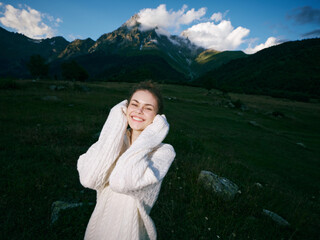 Fototapeta premium Woman, smile, nature, outdoors, mountains, sweater, joy, alpine, landscape, carefree moment in sunny field with towering peaks and blue sky behind her and warm light, fresh air, quiet atmosphere