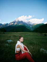 Fototapeta premium Mountain landscape with blue sky and clouds, a woman sits in a meadow wearing red pants and a white top, outdoor fashion in scenic nature.