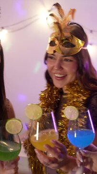 Happy friends in festive costumes dancing with colorful cocktails and smiling at a home party, enjoying the music and celebrating new year's eve together with garlands and vibrant lights