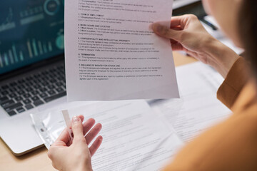 Young adult woman holding contract document while reviewing paperwork at desk, laptop with data charts visible in background, hands and partial profile in focus