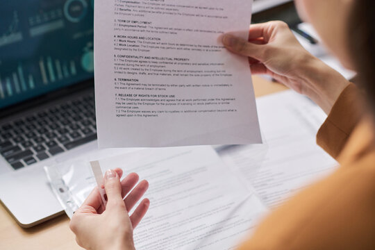 Young adult woman holding contract document while reviewing paperwork at desk, laptop with data charts visible in background, hands and partial profile in focus