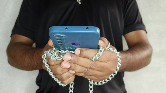 A young adult person hands are bound by chain while holding a smartphone. Mobile phone device addiction, obsession, consumption, imbalance, manipulation, privacy and digital consumerism concept.