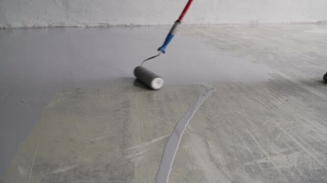 Close-up view of a skilled worker applying a gray epoxy resin coating to a concrete floor using a paint roller for renovation