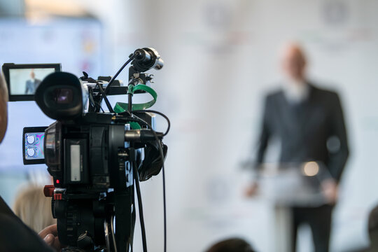 TV camera filming live press event with officials at podium. Highlights media coordination, lighting precision, and structured public communication.