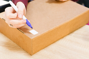 Hand marking white label on cardboard box. Detailed shot of a hand using purple marker to write on a blank tag attached to a brown carton.
