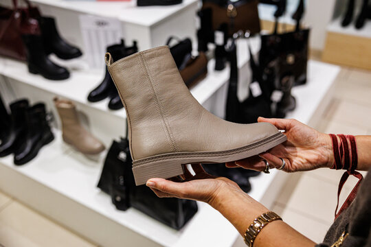 A shopper holds a beige leather ankle boot in a retail store, surrounded by various black and brown boots on display. Close-up shot with indoor lighting