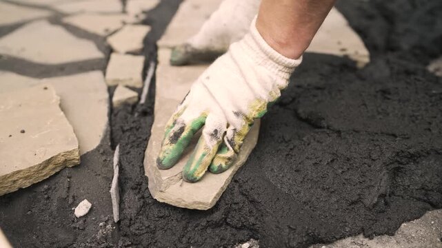 Closeup of a skilled mason wearing protective gloves carefully placing a flagstone paver onto fresh cement for a new garden pathway