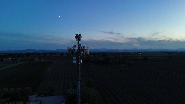 Aerial footage of a tall telecommunication tower equipped with antennas and satellite dishes, transmitting signals against a clear dark blue sky at dusk in a rural landscape