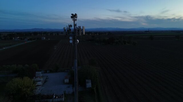 Aerial footage of a tall telecommunication tower equipped with antennas and satellite dishes, transmitting signals against a clear dark blue sky at dusk in a rural landscape