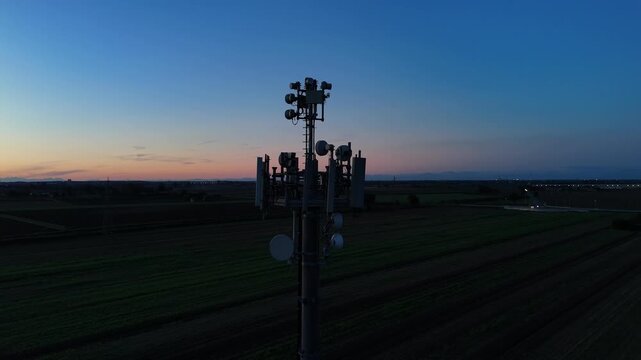 Aerial footage of a tall telecommunication tower equipped with antennas and satellite dishes, transmitting signals against a clear dark blue sky at dusk in a rural landscape
