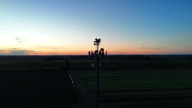Aerial footage of a tall telecommunication tower equipped with antennas and satellite dishes, transmitting signals against a clear dark blue sky at dusk in a rural landscape