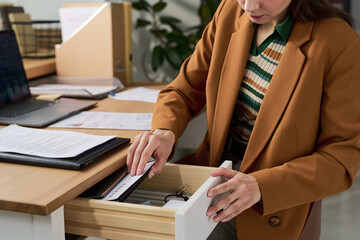 Young adult Caucasian woman opening desk drawer, organizing documents and folders in modern office workspace, focusing on paperwork management and administrative tasks
