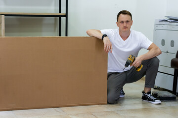 Young man crouching and holding drill next to large cardboard box in office setting