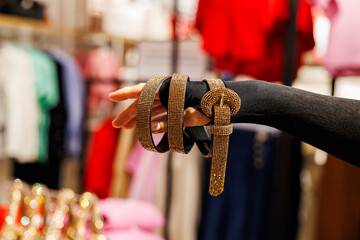 A glittery gold rhinestone-studded belt with a matching round buckle is elegantly draped over a hand in a clothing store with colorful garments in the background