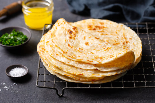 Flaky golden flatbreads cooling on rack, garnished with herbs, salt, and ghee, artisanal food presentation. Dark grey background. Close up.