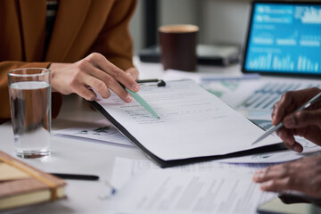 Caucasian woman and Black man collaborating on paperwork at desk, reviewing documents and discussing financial data with laptop and glass of water visible in office setting