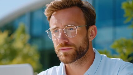 Bearded man in glasses works on a laptop outdoors by a modern office talking on a video call he squints in sunlight, then looks focused as he listens and replies clearly.