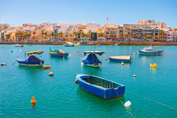 Marsaxlokk Harbour, Malta, is busy with colorful luzzu fishing boats in turquoise waters. Quaint waterfront buildings complete sunny Mediterranean scene
