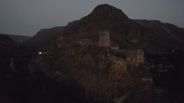 An ancient Khertvisi Fortress illuminated at dusk, standing on a high hill in the Samtskhe Javakheti region of Georgia and overlooking a dramatic mountainous landscape