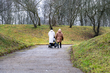 Fototapeta premium Two Women Walking with Baby Stroller Along Park Path in Late Autumn