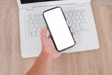 Hand Holding Smartphone With Blank Screen Over Laptop Keyboard on Wooden Desk