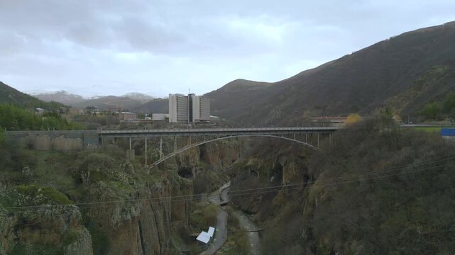 Aerial drone video showcasing a modern bridge over a deep rocky canyon in jermuk armenia with winding river cliffs forested slopes and mountains under soft cloudy sky creating cinematic travel scenery