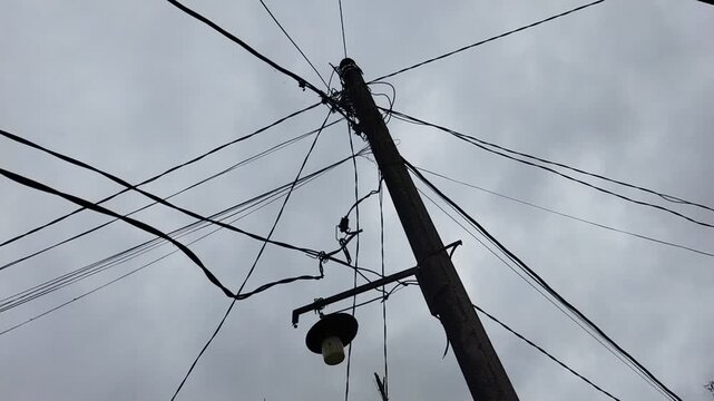 Cinematic low-angle shot of a utility pole with tangled power lines against a moody, overcast sky. A dramatic representation of urban infrastructure and telecommunications under a gloomy atmosphere.