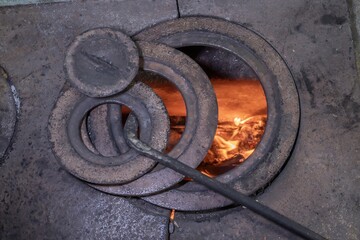 Traditional rural wood fired stove made of stone and metal with open firebox and cast iron rings. Burning logs inside an old village stove showing rustic interior and folk household heritage.