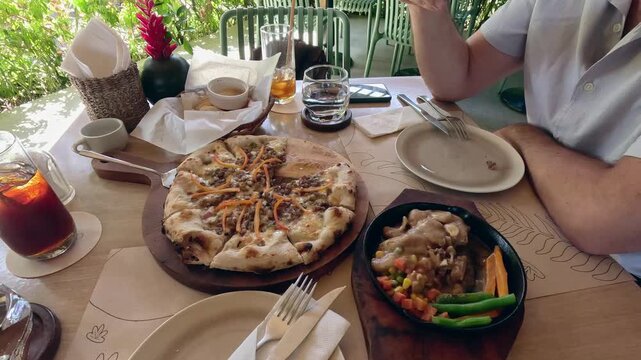 Lunch spread featuring sisig pizza, chicken flambe and drinks on a wooden table in an outdoor dining setup, candid restaurant scene