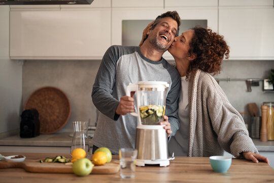 Joyful Couple In A Modern Kitchen Blending Fruit, Sharing A Kiss And Laughing Together