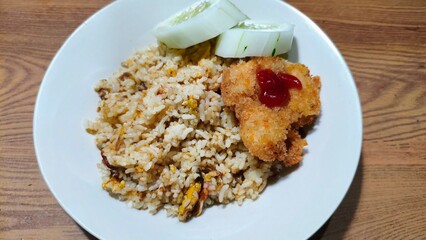 Top view of homemade fried rice with chicken katsu and cucumber on a white plate.