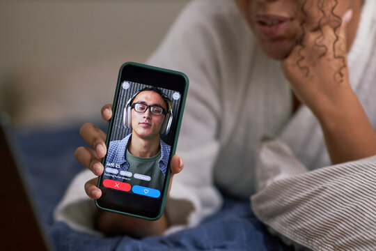 Young adult Black woman holding smartphone showing dating app profile of young Asian wearing glasses, lying on bed, engaging in online dating activity