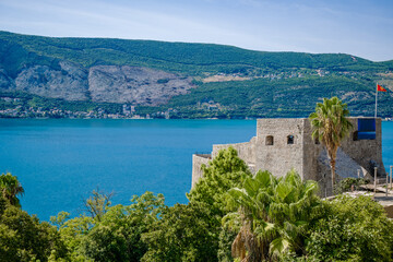 Naklejka premium Scenic view of Herceg Novi, Montenegro, featuring a historic stone fortress by the sea, surrounded by lush greenery and mountains in the background.