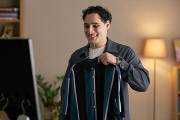 Young adult Caucasian man smiling while holding shirt in front of mirror, preparing for date,...