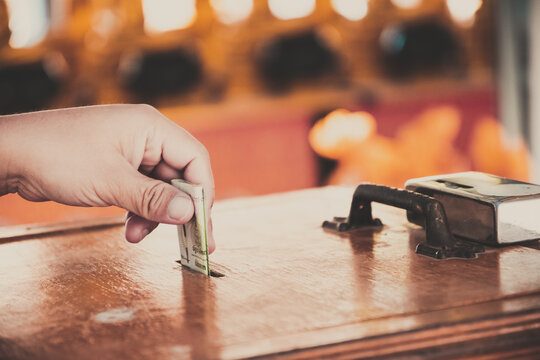 Close-up of a hand putting a Thai Baht banknote into a wooden donation box at a temple. Concept of charity, making merit, generosity, and religious contribution in Southeast Asian culture.