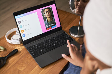 Young adult Black woman sitting at desk using laptop for online dating video call with young adult Asian man, reflection of her face visible in mirror, headphones on table