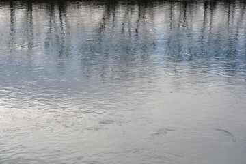 A serene view of a calm river surface reflecting the silhouettes of bare trees and a blue sky