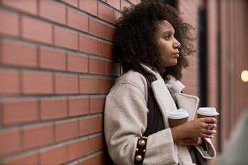 Young adult Black woman standing against brick wall holding two takeaway coffee cups, gazing thoughtfully into distance, suggesting anticipation or waiting in dating context