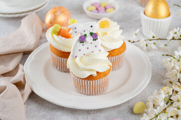 Easter cupcakes are decorated with cream, sugar bunnies, chocolate eggs, jelly carrots on a white plate. Easter food composition. Copy space.