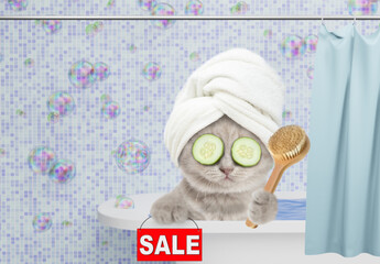 Funny kitten with towel on it head and with slices of cucumber on it eyes takes a bath at home, holds shower brush and shows signboard with labeled "sale". Empty space for text