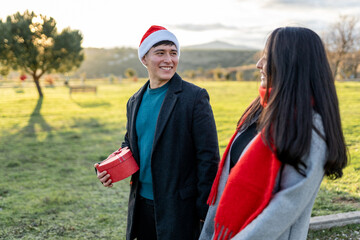 Young man wearing santa hat holding heart-shaped gift box, walking happy with woman in park during christmas holiday