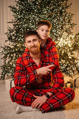 Happy father and young son in matching red plaid pajamas hugging and smiling in front of a glowing Christmas tree at home.