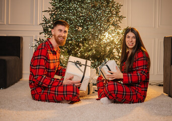 Happy young couple in matching red plaid pajamas holding gift boxes while sitting on a carpet in front of a glowing Christmas tree at home.