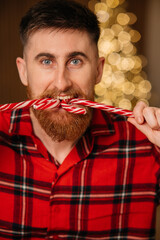 Close-up portrait of a happy bearded man in red plaid pajamas biting traditional Christmas candy canes in front of a blurred glowing tree