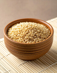 A rustic, handmade clay bowl filled with small, light-colored, raw millet grains is sitting on a clean, simple, light-colored mat.