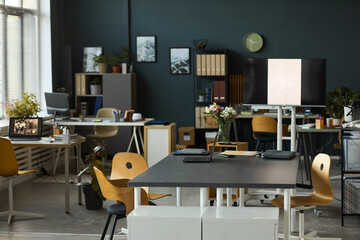 Modern open plan office workspace featuring empty desks with digital tablets and monitors, organized shelves with folders and plants, large windows allowing natural daylight